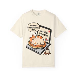 Può includere: T-shirt color crema con un gatto dei cartoni animati che dorme su un laptop. Lo schermo del laptop mostra "Task failed successfully". Un fumetto dice "Not lazy. Energy saving mode". Una tazza di caffè è accanto al laptop.