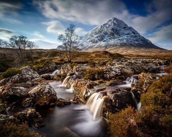 May include: A scenic landscape featuring a snow-capped mountain peak with a cascading waterfall flowing through a rocky stream bed. The sky is a vibrant blue with white clouds.