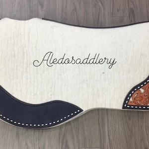 Könnte beinhalten: Ein weißer Sattelpad mit schwarzem Lederbesatz und einem braunen Lederblumenmuster. Das Pad ist mit weißem Garn gestickt und trägt den Text "Aledosaddlery" gestickt.