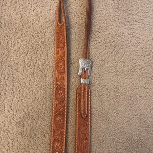 Puede incluir: Una correa de guitarra de cuero marrón con una hebilla plateada y tallado floral. La correa está hecha de dos piezas de cuero cosidas juntas.