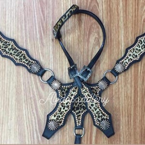 May include: A black leather horse bridle with a leopard print design and silver accents. The bridle includes a headstall, reins, and a breast collar.