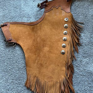 May include: Brown leather chaps with fringe and silver buttons. The chaps have a leather strap with a buckle on the top and the text "S.M. Frank Saddlery Co." is written on the back.