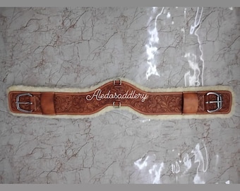 May include: A brown leather cinch with a white sheepskin lining and silver buckles. The cinch is decorated with a floral tooled design and the text "Aledosaddlery".