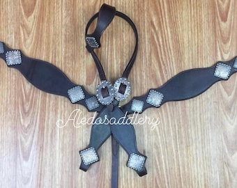 Puede incluir: Una cabezada de caballo de cuero marrón con conchos plateados y un pecho de caballo a juego. La cabezada tiene un frontal, un bozal y riendas. El pecho de caballo tiene una correa de pecho y dos correas laterales.