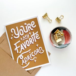 May include: A mustard yellow card with white lettering that reads "You're my favourite Capricorn". The card is surrounded by a brown envelope and gold binder clips.