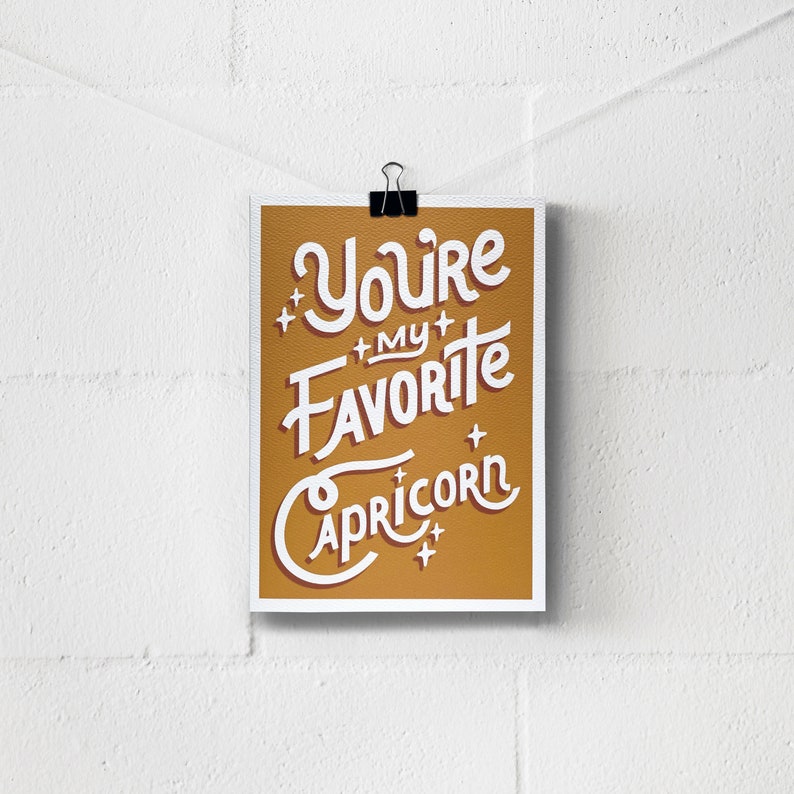 May include: A gold-coloured print with white text that reads "You're my favourite Capricorn" with white stars around the text.