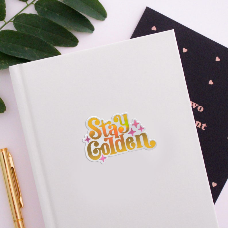 May include: A gold sticker with the text "Stay Golden" in a retro font. The text is in a gradient of yellow, orange, and pink. There are small pink stars around the text.
