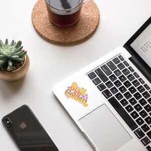 May include: A laptop computer with a sticker that says "Stay Golden" in a rainbow gradient font with pink stars. The sticker is on the silver laptop case, above the keyboard.