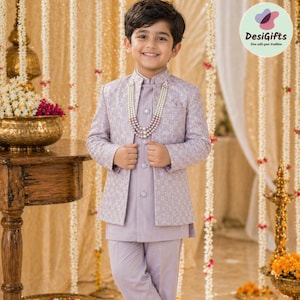 May include: A young boy in a lavender suit with a patterned jacket and matching pants. He wears a pearl necklace and black dress shoes. The background features floral decorations and the text "DesiGifts: Live with your tradition."