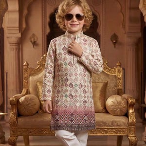 May include: A young person wearing a cream-colored kurta with a geometric pattern in shades of pink, green, and purple, paired with white pants. The outfit is completed with patterned shoes and sunglasses. The setting appears to be an ornate room with gold accents.