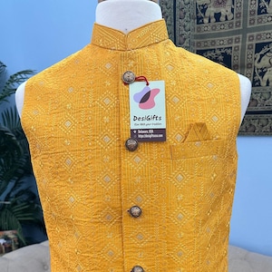May include: Mustard yellow vest with a geometric pattern and decorative buttons. The vest has a mandarin collar and a pocket on the chest. A tag from DesiGifts is attached. The vest is displayed on a mannequin.