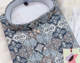 Può includere: Una camicia abbottonata fantasia con colletto alla coreana. La camicia presenta un design geometrico nei toni del blu, marrone e bianco. La camicia ha un'etichetta con la scritta "DesiGifts".