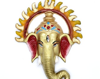 May include: A gold-colored Ganesha head with a red and white painted face, a third eye, and a blue and red crown. The head is surrounded by a yellow and orange halo.