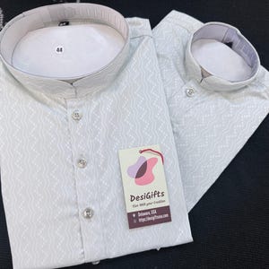 May include: A light gray long-sleeved shirt with a mandarin collar and a subtle white zigzag pattern. The shirt features a front button placket and a size 44 tag inside the collar. A DesiGifts brand tag is attached to the front of the garment.