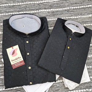 May include: Two black kurta shirts with gold buttons and white collars. The shirts are made of a textured fabric and have a traditional Indian design. The shirts are folded and placed on a white surface. The text "DesiGifts" is visible on a tag attached to one of the shirts.