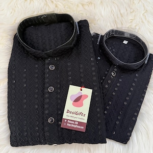 May include: Two black embroidered kurta shirts with a mandarin collar and button-up front. The shirts have a detailed pattern and are displayed on a white, fluffy surface. A tag with the text "DesiGifts" is attached.