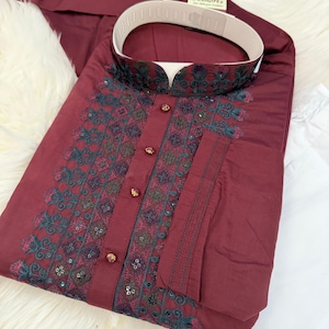 May include: A maroon kurta with a white collar and intricate black and green embroidery. The embroidery features sequins and a floral design. The kurta is folded and laid on a white fluffy surface. The text "Desigifts" is visible on a small tag.