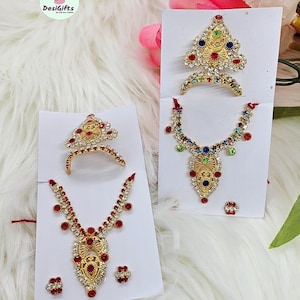 May include: Two sets of gold-tone jewelry with red, blue, and green gemstones. The jewelry includes a crown, necklace, and earrings. The text "Design -1" is printed on the bottom of the image.