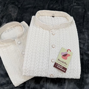 May include: Two white, embroidered kurta shirts are neatly folded and displayed. The shirts have a mandarin collar and button-down front. A tag with the text "DesiGifts" is attached to one shirt.
