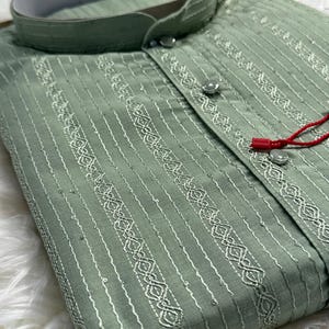 May include: Sage green kurta shirt with vertical white embroidered stripes and decorative patterns. The shirt features a mandarin collar, button-down front, and a red string detail. The fabric has a subtle sheen.