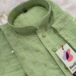May include: A light green, embroidered kurta with a mandarin collar and decorative buttons. The fabric features a detailed pattern of floral and geometric designs. A tag with the text "DesiGi" is attached to the garment.