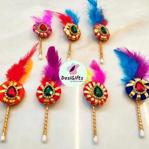 May include: A collection of colorful, decorative feather brooches. Each brooch features a jewel-encrusted, circular centerpiece in various colors, with a gold stem and a cluster of vibrant feathers. The brooches are arranged on a light surface.