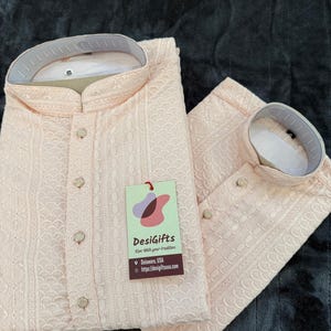 May include: Two light peach-colored kurta shirts with intricate embroidered patterns and button closures. The shirts are neatly folded on a dark gray textured surface. A tag with the text "DesiGifts" is attached to one of the shirts.