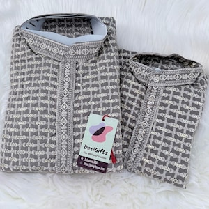 May include: Two gray embroidered kurta shirts with white accents, featuring intricate geometric and floral patterns. The shirts are folded and displayed on a white, fluffy surface. A tag with the text "DesiGifts" is attached.
