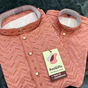 May include: Two coral-colored, embroidered shirts with a mandarin collar and gold-colored buttons. The shirts have a chevron pattern and a tag that reads "DesiGifts". The shirts are likely part of a traditional clothing set.