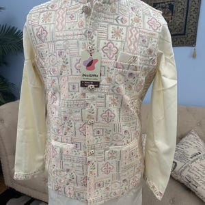 May include: Cream-colored embroidered vest with a floral and geometric pattern, paired with a matching long-sleeved shirt and pants. The vest features a mandarin collar and decorative buttons. A tag with the text "DesiGifts" is attached.