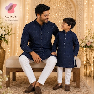 May include: A father and son are seen wearing matching navy blue embroidered kurta shirts with white pants. The father is seated, and the son is standing, both smiling. The image is set in a warmly lit room with floral arrangements.