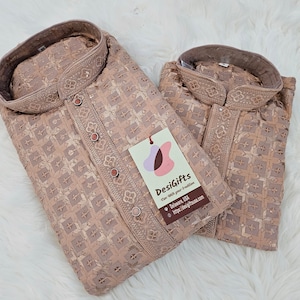 May include: Two beige embroidered kurta shirts with a mandarin collar and button-down front. The shirts have a detailed geometric pattern and a tag that reads "DesiGifts". The shirts are displayed on a white, fluffy surface.