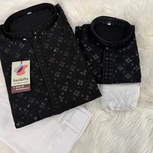 May include: Two black kurta shirts with white embroidery and a white label that says "Desigifts" on the larger shirt. The shirts are on a white fluffy surface.