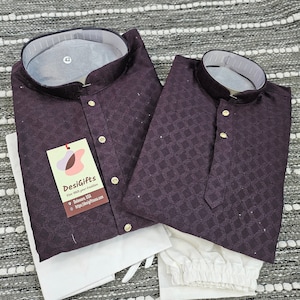 May include: Two purple patterned shirts with white collars and buttons. The shirts are folded and laid out on a white and gray patterned surface. A white tag with the text "DesiGifts" is attached to the larger shirt. The tag also includes the text "For Dress Your Tradition" and a website address.