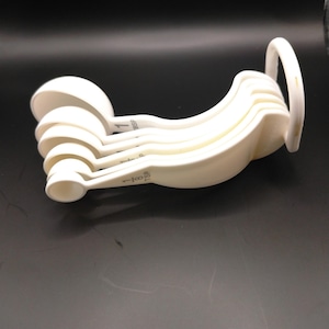 Può includere: Un set di cinque misurini in plastica bianca impilati su un anello in plastica bianca. I misurini sono contrassegnati con le misure 1 TBSP, 1/2 TBSP, 1 TSP, 1/2 TSP e 1/8 TSP, con il testo chiaramente stampato sui manici.