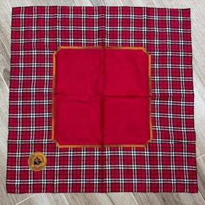 May include: A red and white tartan scarf with a solid red central panel. The panel is framed with a gold border. A gold emblem is in the corner of the scarf.