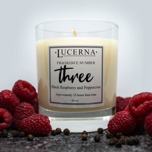 Może przedstawiać: Szklany słoik na świecę z przezroczystego szkła z białą etykietą, na której widnieje napis "·LUCERNA· FRAGRANCE NUMBER three Black Raspberry and Peppercorn Approximatly 35 hours burn time 10 oz". Świeca jest otoczona malinami i czarnymi ziarenkami pieprzu.