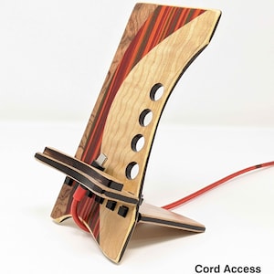 May include: A wooden phone stand with a red, orange and yellow striped design. The stand has a cutout for charging cables and a slot for holding a phone. The stand is made of wood and has a natural finish. "Cord Access" is written in black text at the bottom of the image.
