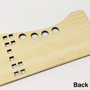 May include: A light brown wooden tool with a curved top and a flat bottom. The tool has several round holes and square holes cut out of it. The back side of the tool is shown in the image.
