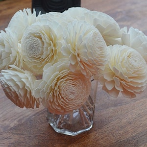 May include: A bouquet of white sola wood flowers in a clear glass vase. The flowers are arranged in a circular pattern and have a delicate, layered texture.