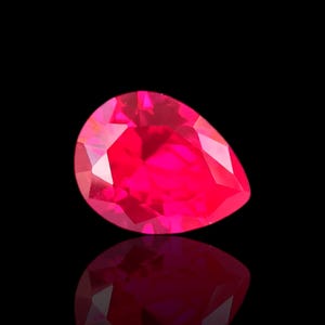May include: A faceted, pear-shaped ruby gemstone, radiating a vibrant red hue. The gem is set against a black background, with its reflection visible below. The cut of the stone creates multiple facets that reflect light.