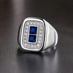 May include: A silver-toned ring featuring a rectangular face with two stacked, square-cut deep blue stones at the center. The blue stones are surrounded by a border of small, clear, round-cut stones set into the polished metal frame of the ring.