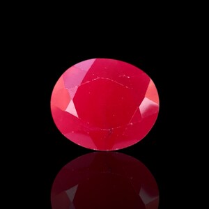 May include: A faceted, round, bright red gemstone against a black background. The gem has a high degree of clarity and reflects light, creating a brilliant appearance. The gem's reflection is visible below it.