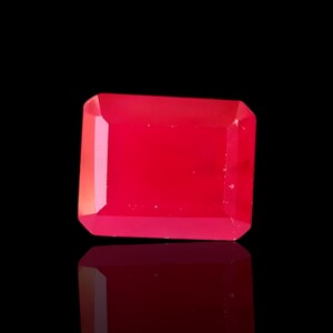 May include: A vivid, rectangular-cut, red gemstone with a polished surface. The gem is set against a black background, with its reflection visible below. The gemstone's facets catch the light, creating a brilliant display.