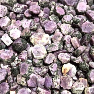 May include: A close-up view of a pile of rough, purple gemstones. The stones are various sizes and shapes, with a rough, natural finish.