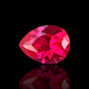 Puede incluir: Una gema de rubí facetada en forma de pera, que irradia un tono rojo vibrante. La gema se encuentra sobre un fondo negro, con su reflejo visible debajo. El corte de la piedra crea múltiples facetas que captan y reflejan la luz.