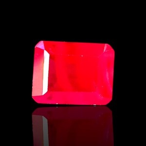 Puede incluir: Una vibrante gema de rubí de corte rectangular, que irradia un tono rojo intenso. Las facetas de la gema capturan la luz, creando reflejos brillantes sobre un fondo negro. La superficie pulida de la piedra refleja la luz, realzando su brillo.
