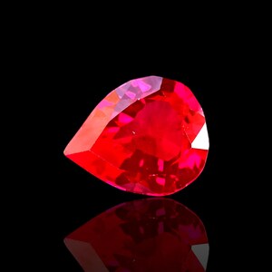 May include: A faceted, pear-shaped, vibrant red gemstone. The gem has a high degree of clarity and brilliance, reflecting light to create a dazzling display. The gemstone is set against a black background, with its reflection visible below.