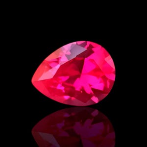 Op de afbeelding: Een gefacetteerde, peervormige edelsteen in een levendige tint roze. De edelsteen is geplaatst tegen een zwarte achtergrond, met zijn weerspiegeling eronder zichtbaar. De slijping van de steen creëert meerdere facetten die licht reflecteren.