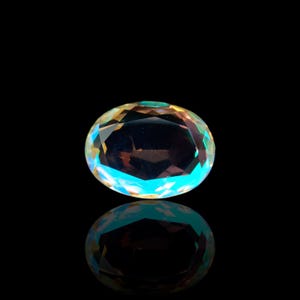 May include: An oval-cut gemstone with a rainbow iridescence, displaying hues of yellow, blue, and green. The gem is set against a black background, with its reflection visible below.